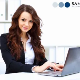 Pracownica w biurze w Warszawie, w czarnej marynarce i białej koszuli, uśmiecha się i pracuje na laptopie; w tle białe regały z segregatorami.