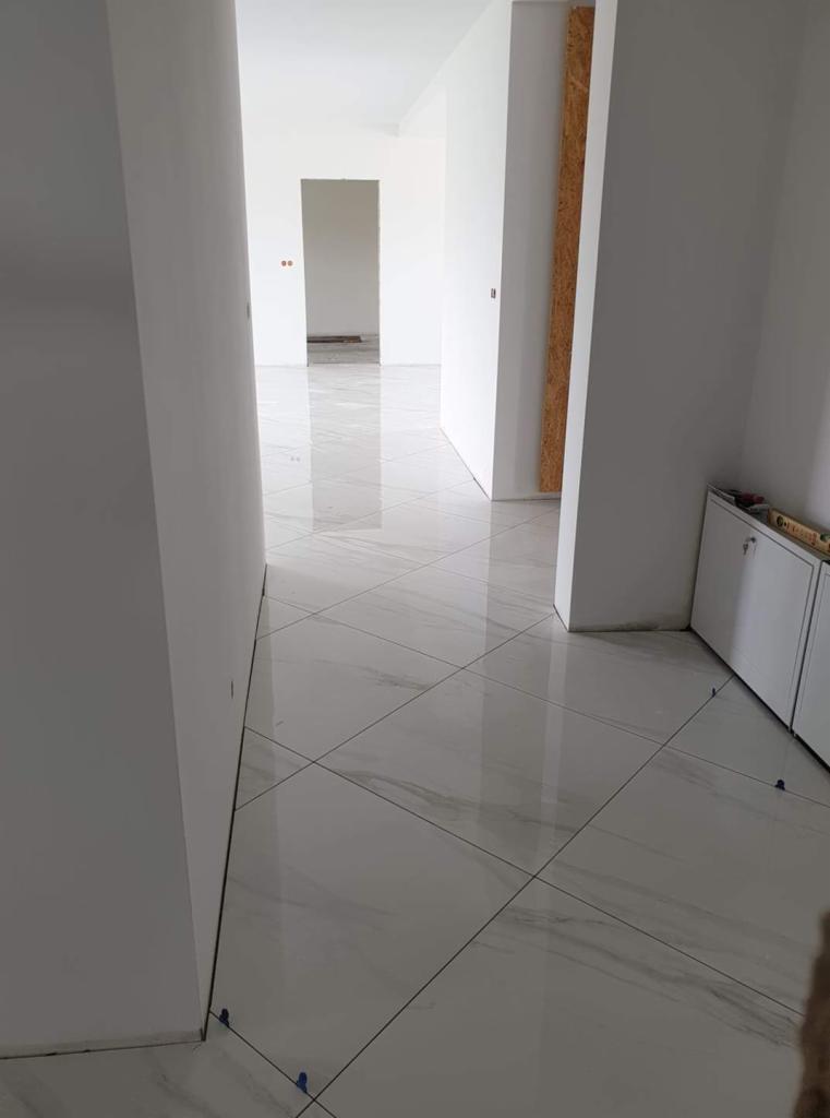 Białe płytki podłogowe z szarym żyłkowaniem ułożone diagonalnie w korytarzu podczas prac wykończeniowych, widoczne krzyżyki dystansowe, świeżo pomalowane ściany na biało.