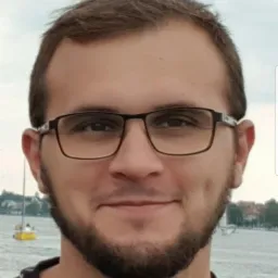 Portret uśmiechniętego mężczyzny w okularach na tle jeziora z żaglówką, ubrany w koszulkę z motywem koniczyny i podkowy. Ujęcie z bliska, lekki uśmiech.