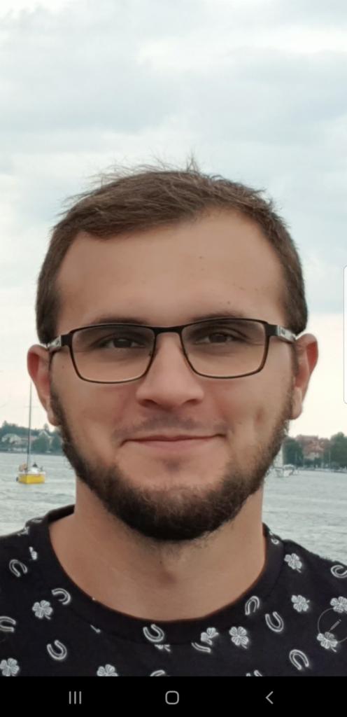 Portret uśmiechniętego mężczyzny w okularach na tle jeziora z żaglówką, ubrany w koszulkę z motywem koniczyny i podkowy. Ujęcie z bliska, lekki uśmiech.