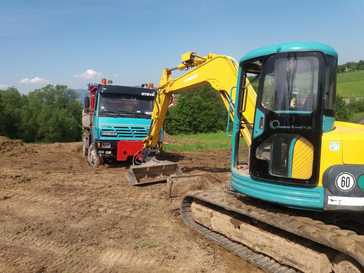 Żółto-turkusowa koparka Yanmar Round Cab i błękitna ciężarówka Steyr na placu budowy, prawdopodobnie podczas prac ziemnych, z widocznym wykopanym terenem i zielonymi wzgórzami w tle.
