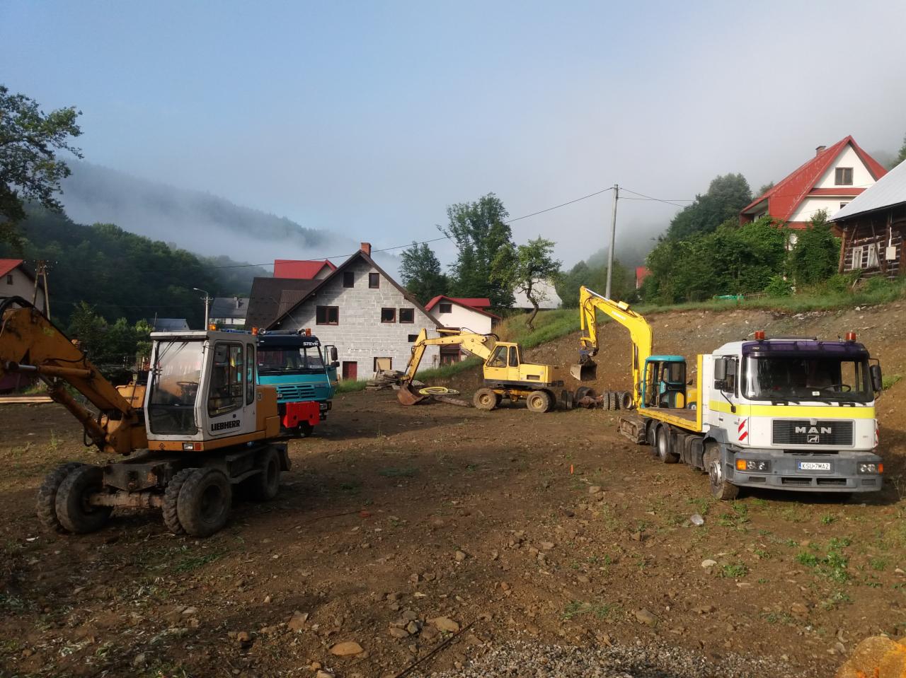 Dwa żółte baggery Liebherr i ciężarówka MAN na placu budowy w pagórkowatym terenie; w tle domy i mgła.