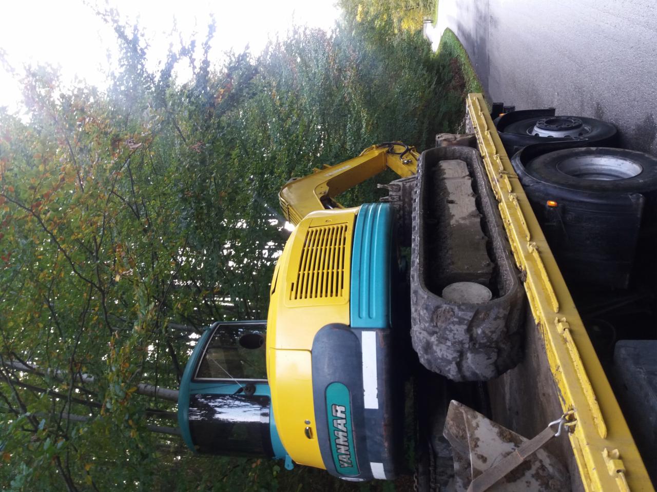 Żółto-turkusowa minikoparka Yanmar przewożona na żółtej lawecie, widok z boku na tle drzew.