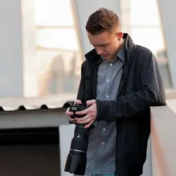 Młody fotograf z aparatem i obiektywem w dłoniach, pochylony nad sprzętem, w tle jasne, rozmyte okno. Ujęcie z lekkiej góry, widoczny kolczyk w uchu.