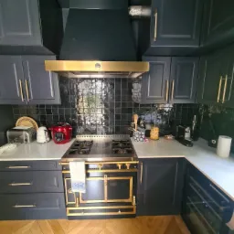 Elegancka kuchnia w odcieniach granatu i złota: zabudowa z ciemnymi szafkami, złote uchwyty, luksusowy piec z elementami złota, biały blat, czarne płytki na ścianie i parkiet w jodełkę na podłodze.