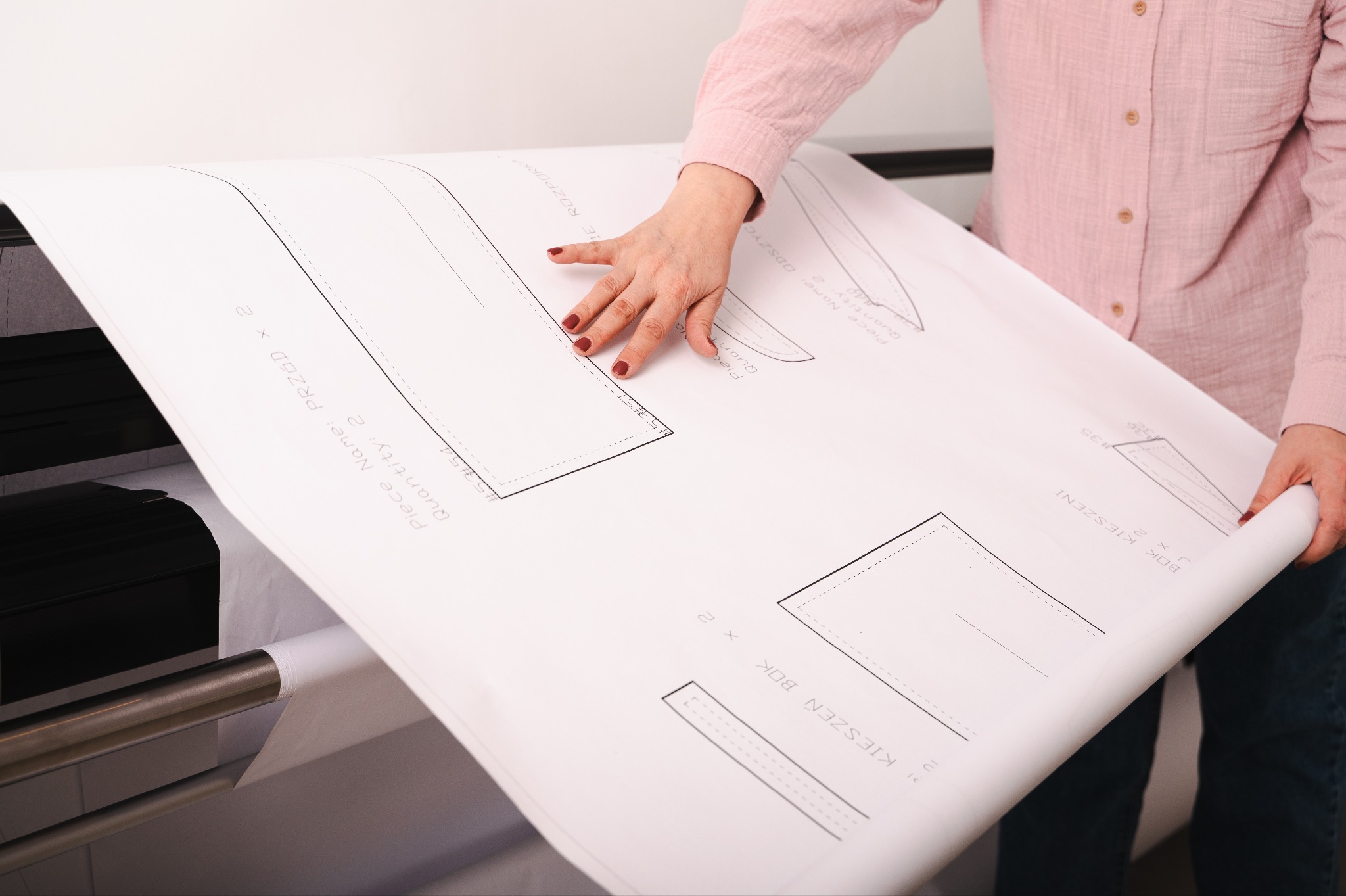 Kobieta w różowej koszuli rozkłada duży arkusz z szablonami odzieżowymi wydrukowanymi na drukarce. Widoczne napisy: 'PRZÓD', 'KIESZEŃ BOK'.