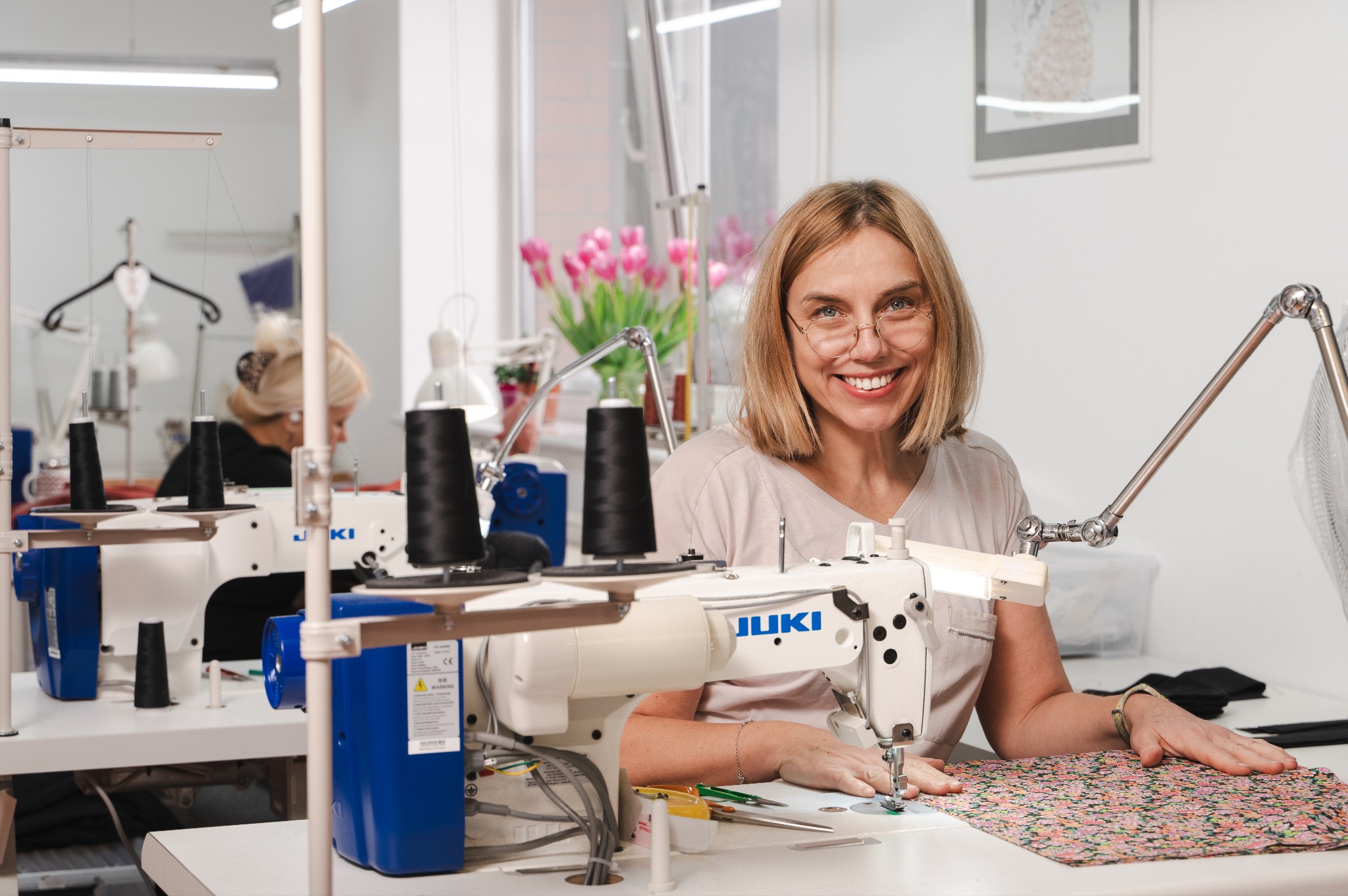 Uśmiechnięta blondynka w okularach szyje na maszynie JUKI w jasnej szwalni. W tle tulipany i druga maszyna. Profesjonalne oświetlenie i schludne stanowisko pracy.