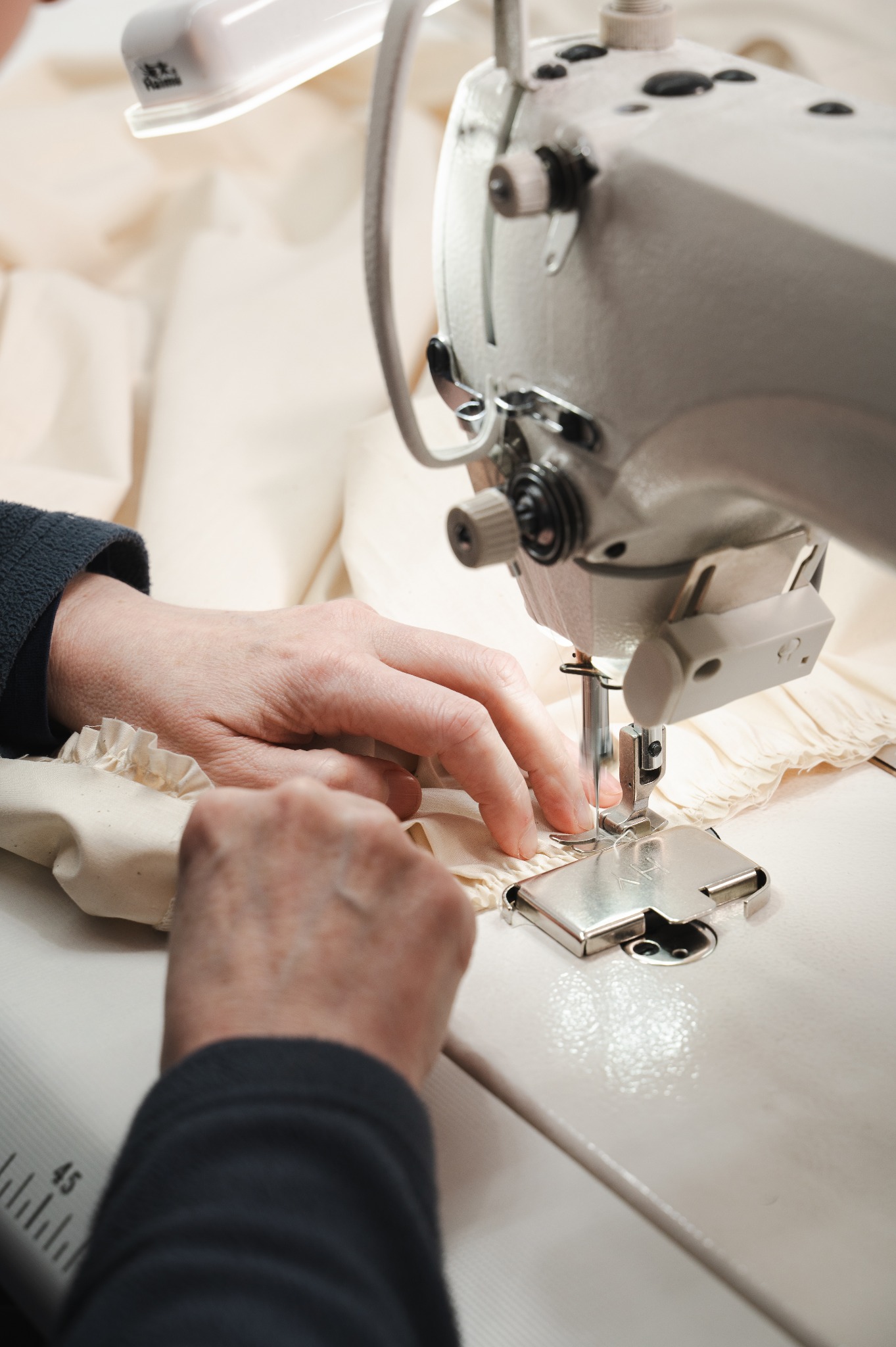 Detal szycia na maszynie przemysłowej jasnej tkaniny z falbanką, dłonie krawcowej w trakcie pracy, oświetlenie robocze maszyny.