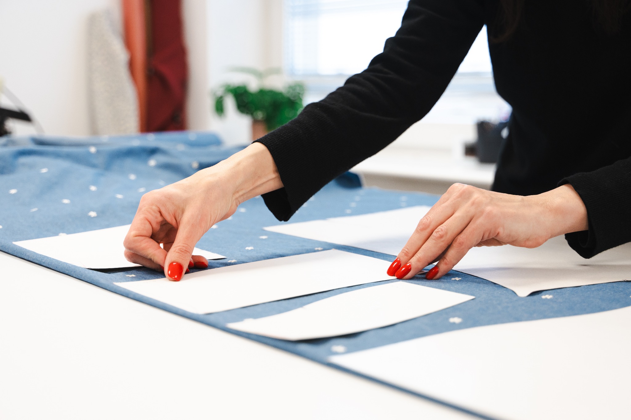 Krawcowa z czerwonym manicure układa białe szablony na błękitnym materiale w białe groszki na białym stole, przygotowując wykroje do szycia odzieży.