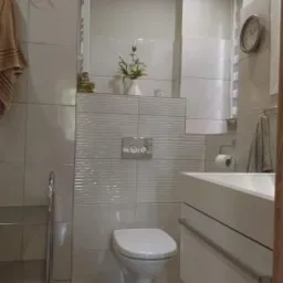 Jasna łazienka z toaletą wiszącą, umywalką w zabudowie, grzejnikiem drabinkowym i zegarem na ścianie.