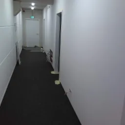 Korytarz w trakcie wykańczania, widoczne świeżo pomalowane ściany, czarna wykładzina, wystające kable i zabezpieczenia taśmą malarską wokół ościeżnic i gniazdek.