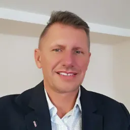 Portret uśmiechniętego mężczyzny w ciemnej marynarce z przypinką w kolorach flagi, na jasnym tle.