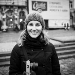 Portret uśmiechniętej kobiety z aparatem fotograficznym na tle miejskiego placu i zabytkowej architektury w czerni i bieli.