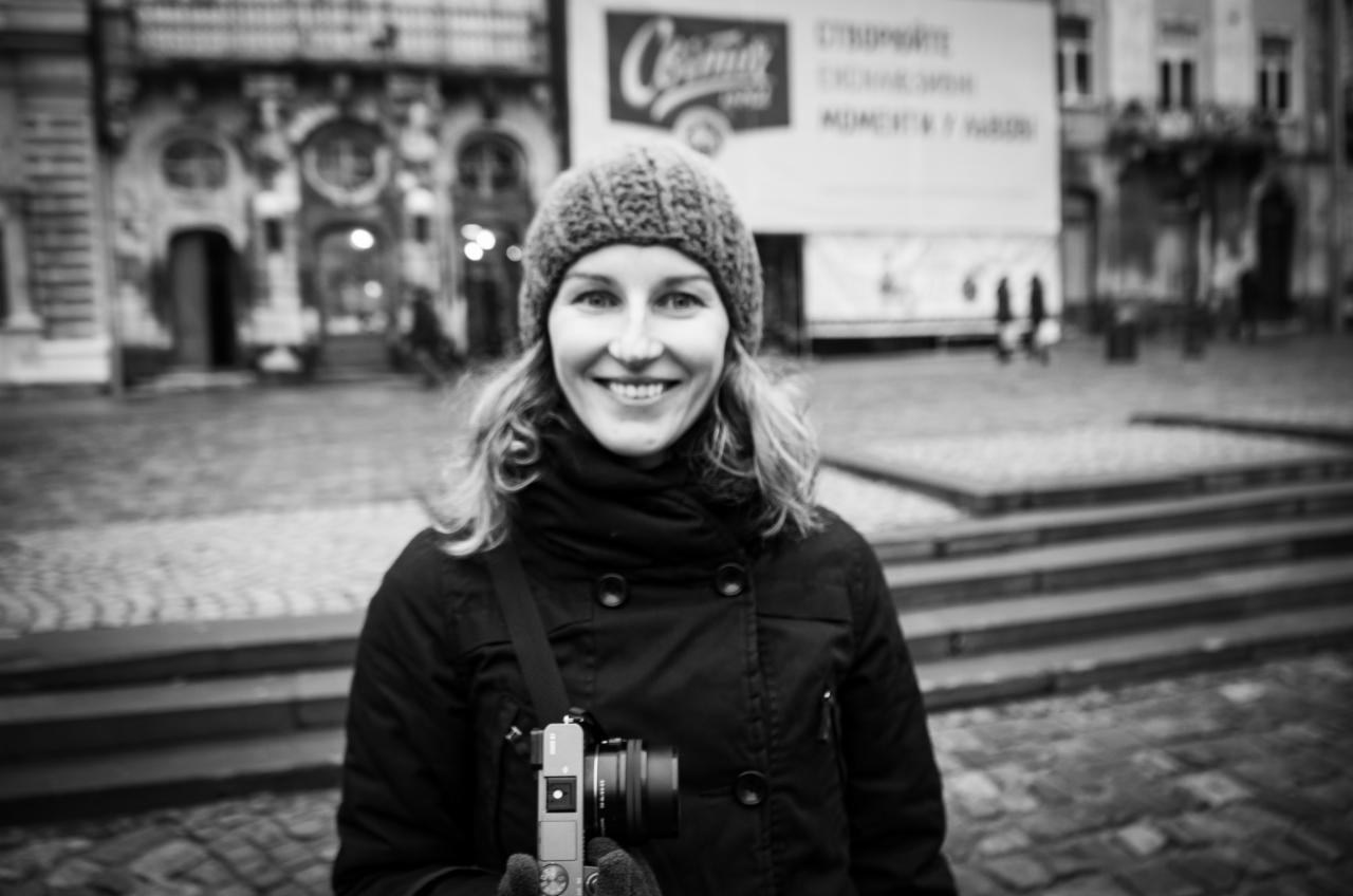 Portret uśmiechniętej kobiety z aparatem fotograficznym na tle miejskiego placu i zabytkowej architektury w czerni i bieli.