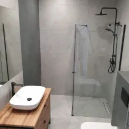 Nowoczesna łazienka z szarymi płytkami, kabiną prysznicową typu walk-in z czarnymi detalami, umywalką nablatową na drewnianej szafce i toaletą.