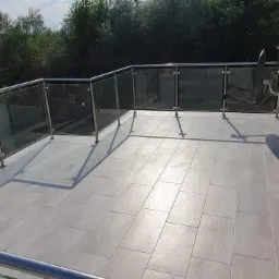 Jasnoszary taras wyłożony prostokątnymi płytkami, zabezpieczony szklaną balustradą ze stali nierdzewnej, z widoczną anteną satelitarną na ścianie budynku.