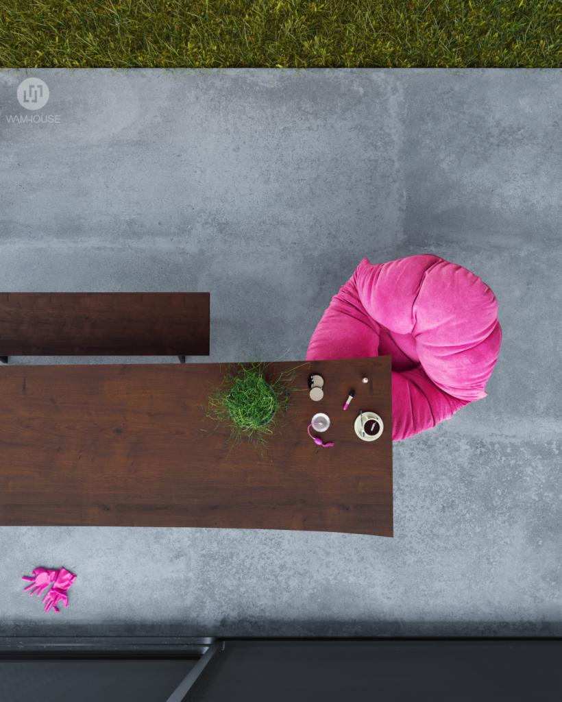 Widok z góry na stół z ciemnego drewna, różowy puf, doniczkę z trawą i akcesoria kosmetyczne na szarym tarasie graniczącym z trawnikiem.