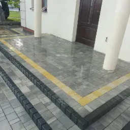 Schody wejściowe wyłożone szarą kostką z czarnymi obrzeżami i żółtymi akcentami, widoczne kolumny podtrzymujące zadaszenie i częściowo zaparkowany samochód w tle.