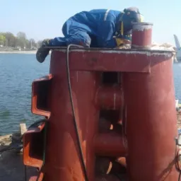 Pracownik w kombinezonie, z maską spawalniczą, klęczy na konstrukcji pomalowanej na czerwono, prawdopodobnie podczas prac spawalniczych na nabrzeżu.