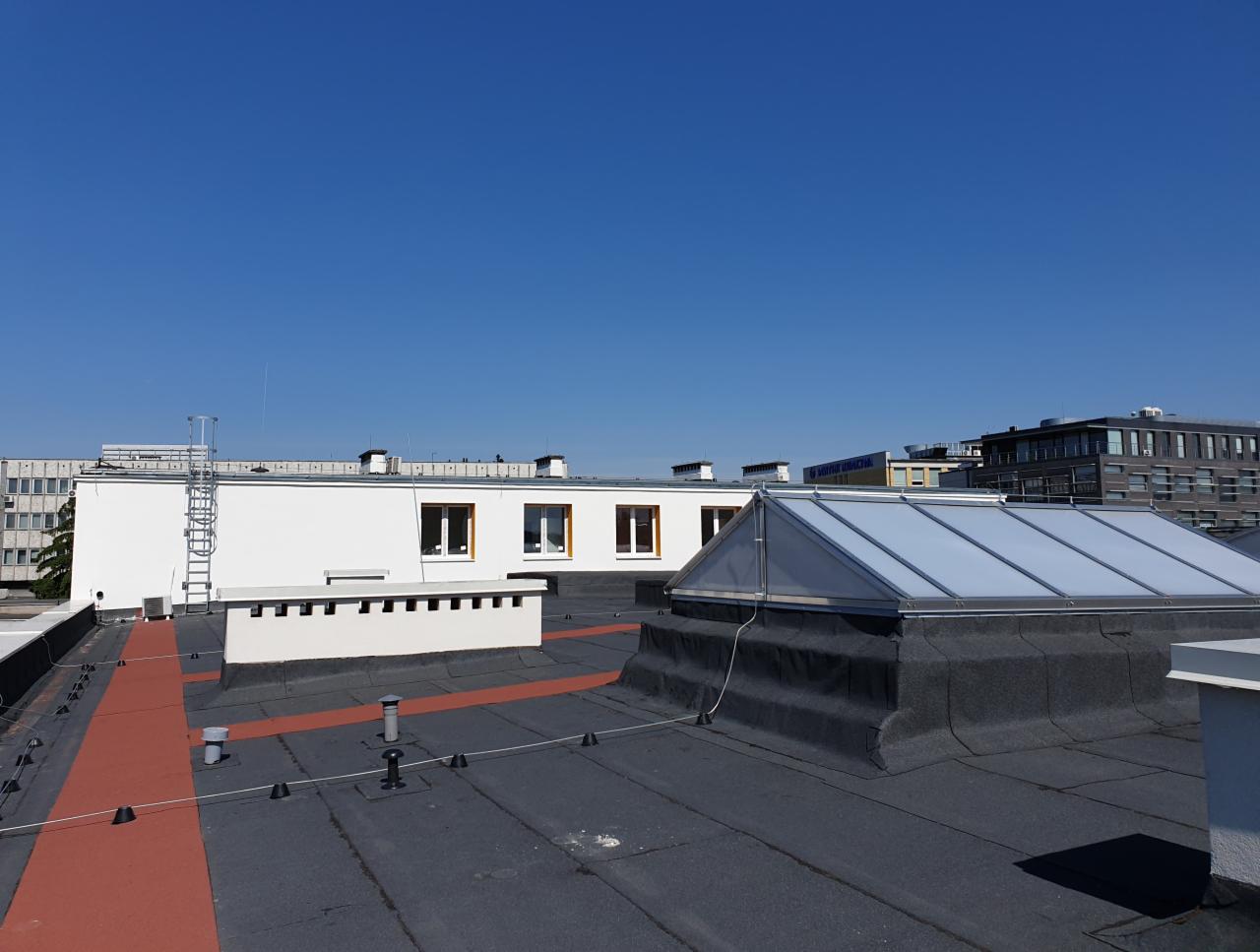 Widok z góry na płaski dach budynku z zainstalowanym świetlikiem dachowym i kominem wentylacyjnym, widoczne również inne budynki w tle pod błękitnym niebem.
