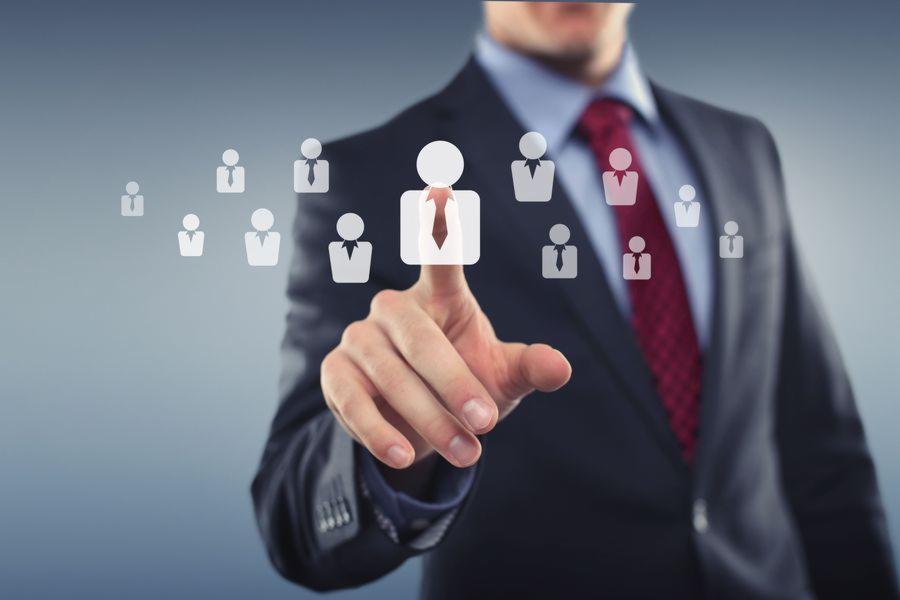 Mężczyzna w garniturze dotyka wirtualnego interfejsu z ikonami ludzi, z naciskiem na ikonę z czerwoną krawatką, symbolizującą wybór pracownika.