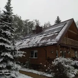 Drewniany dom jednorodzinny z panelami słonecznymi na dachu, częściowo pokrytym śniegiem, otoczony ośnieżonymi drzewami iglastymi i krzewami w zimowej scenerii.