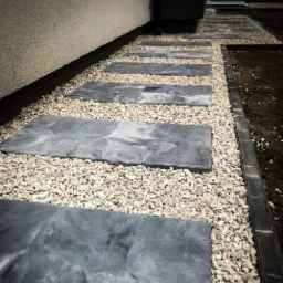 Ścieżka z szarych płyt imitujących kamień, oddzielonych drobnym, jasnym żwirem, prowadząca wzdłuż ściany budynku. Widoczne ciemne obrzeże z kostki po prawej stronie.