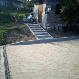 Ułożona kostka brukowa w odcieniach beżu i brązu z widocznym odpływem liniowym, prowadząca do betonowych schodów i murowanej budowli w tle.