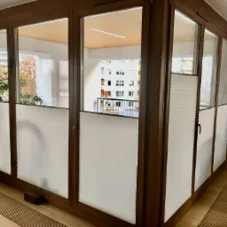 Białe, harmonijkowe osłony okienne zamontowane w brązowych ramach okiennych na zabudowanym balkonie z widokiem na budynek mieszkalny i drzewa.