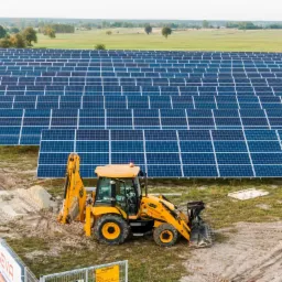 Żółta koparko-ładowarka na tle rozległej farmy paneli słonecznych w słoneczny dzień, z widocznymi śladami opon i stertą piasku obok.