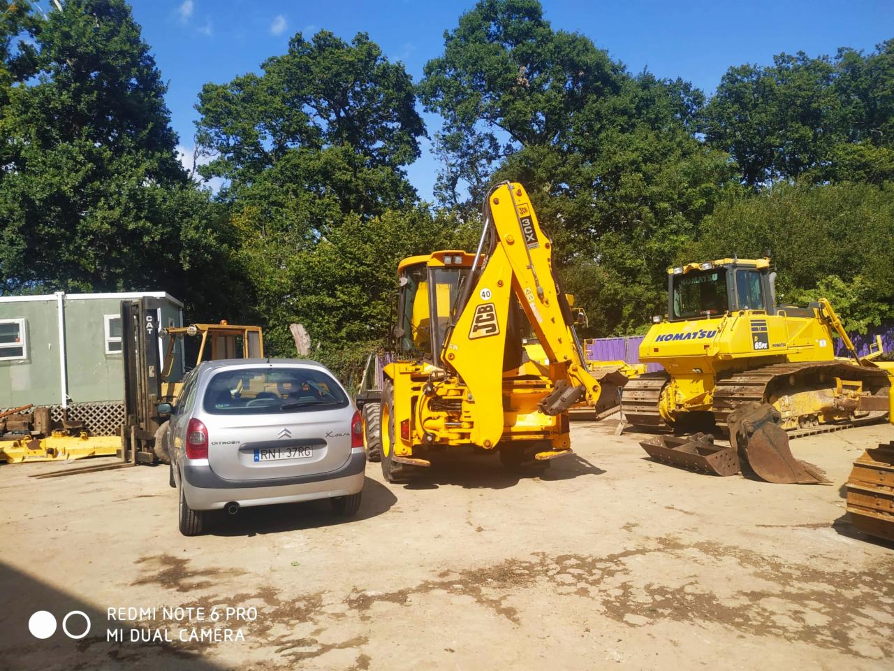 Plac z zaparkowanym srebrnym Citroenem Xsara, obok żółta koparko-ładowarka JCB, spychacz Komatsu i wózek widłowy, w tle zielone drzewa.
