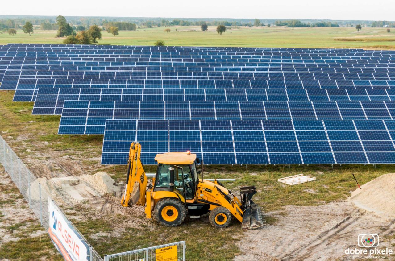 Żółta koparko-ładowarka na tle rozległej farmy paneli słonecznych, widok z góry, częściowo ogrodzony teren budowy.