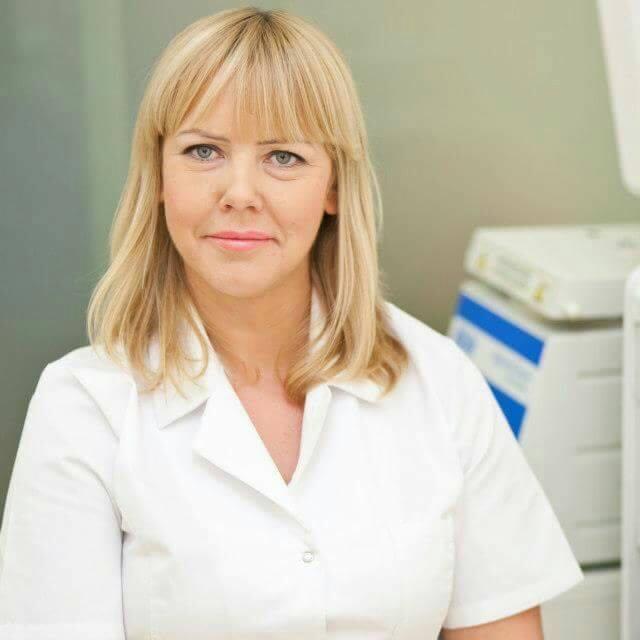 Portret blondynki w białym uniformie na tle gabinetu kosmetycznego. Widoczne profesjonalne urządzenia do zabiegów. Uśmiechnięta twarz, jasne oświetlenie.