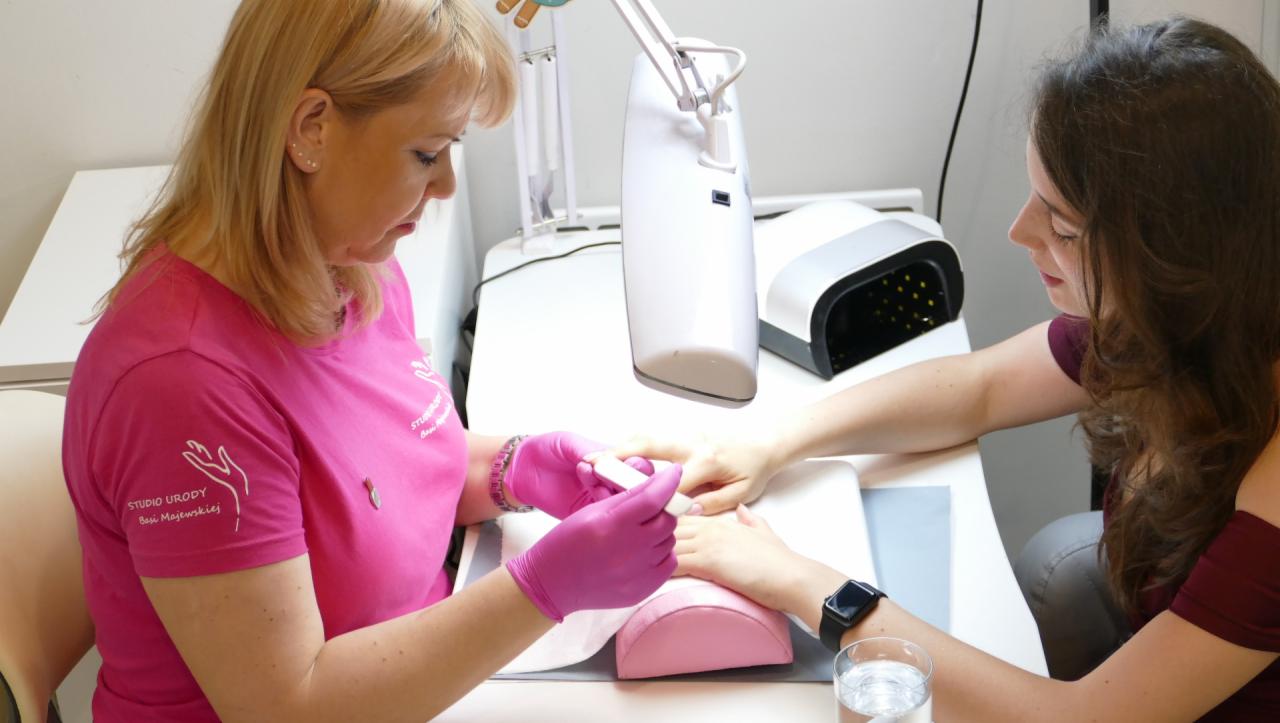 Stylistka w różowych rękawiczkach opracowuje paznokcie klientki pilnikiem w jasnym salonie, widoczna lampa UV i logo studia na koszulce stylistki.