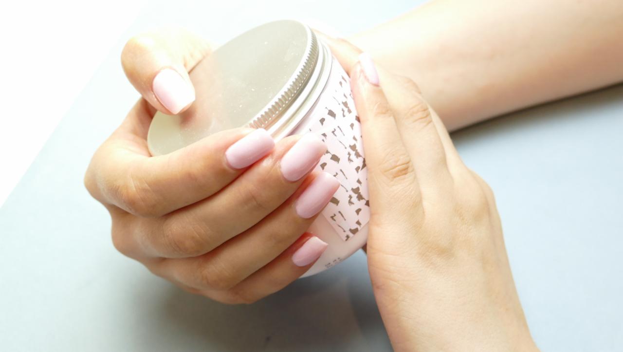 Ręce kobiety z delikatnym, pastelowym różowym manicurem trzymają słoik z kremem o podobnej kolorystyce, na jasnym tle.