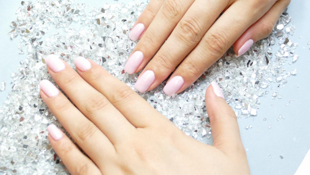 Dłonie z pastelowymi, różowymi paznokciami na tle rozsypanych, srebrnych, błyszczących elementów dekoracyjnych.