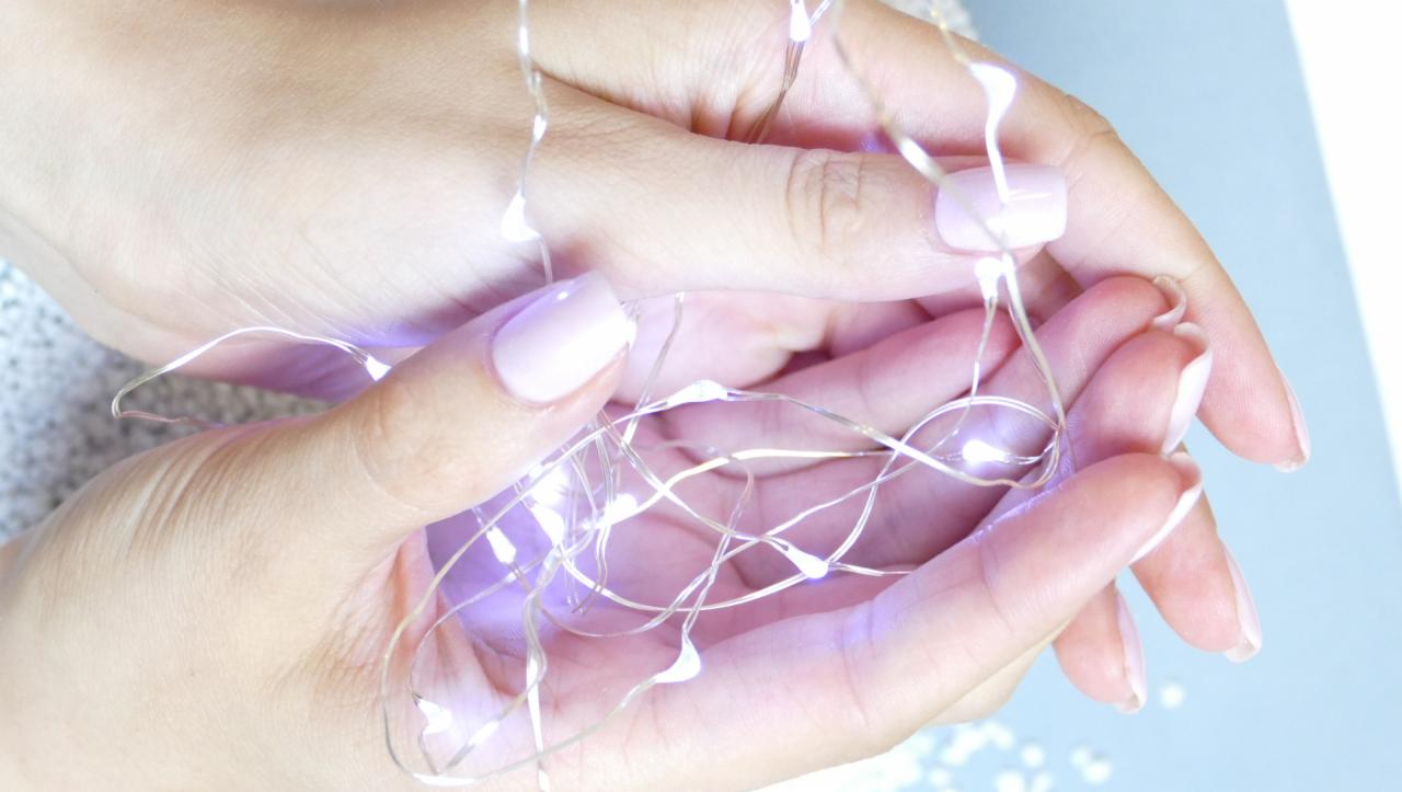 Dłonie kobiety z delikatnym, pastelowym manicure hybrydowym, trzymające delikatny łańcuch światełek LED, tworzący subtelny, świąteczny nastrój.