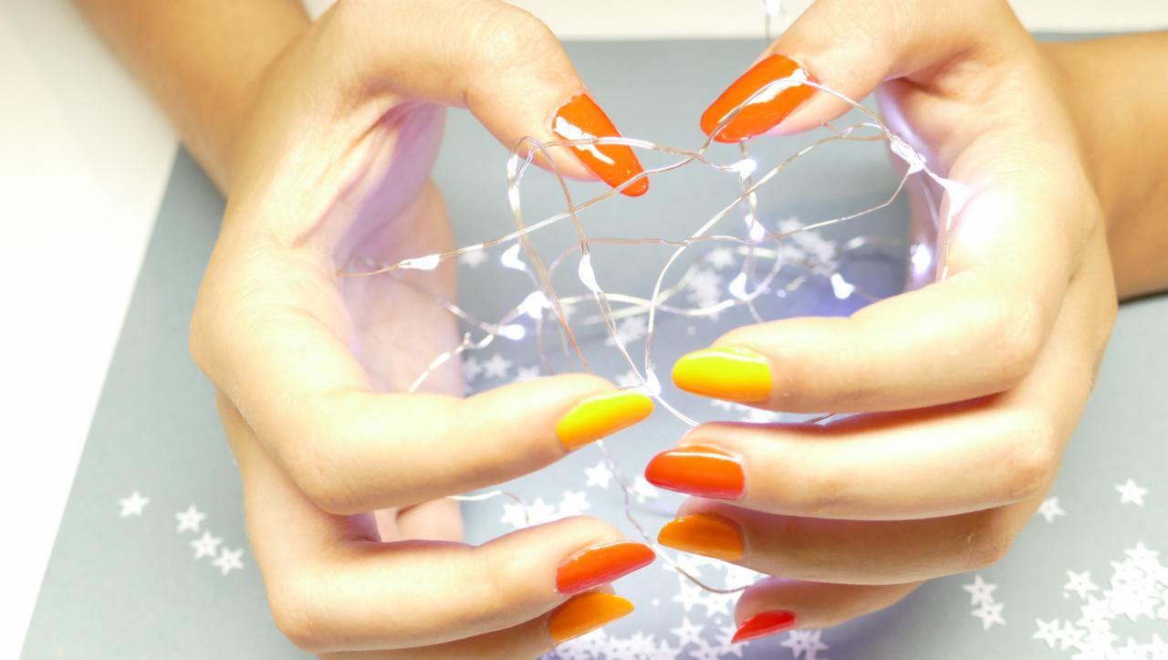 Dłonie z gradientowym pomarańczowo-żółtym lakierem do paznokci trzymają delikatne światełka na tle szarej podkładki ze wzorem gwiazdek.