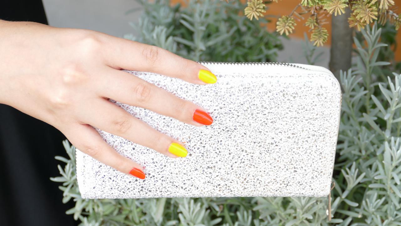 Dłoń z naprzemiennie pomalowanymi paznokciami na żółto i pomarańczowo, trzymająca biało-srebrną, teksturowaną torebkę na tle zielonej roślinności.