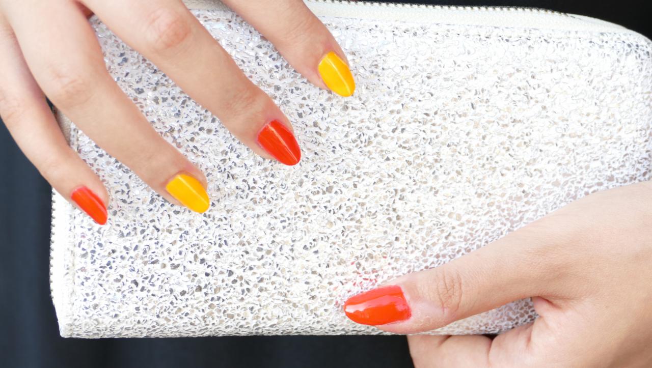 Dłoń z pomalowanymi na przemian żółtym i pomarańczowym lakierem paznokciami, trzymająca srebrną, błyszczącą kopertówkę.