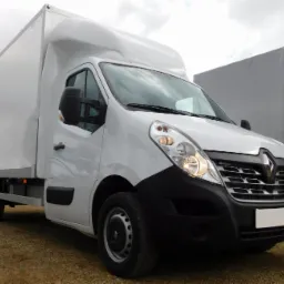 Biały Renault Master z zabudową kontenerową na placu, w tle inne busy i ciężar&oacute;wki. Widok z przodu, lekko z boku. Światło dzienne, pochmurne niebo.