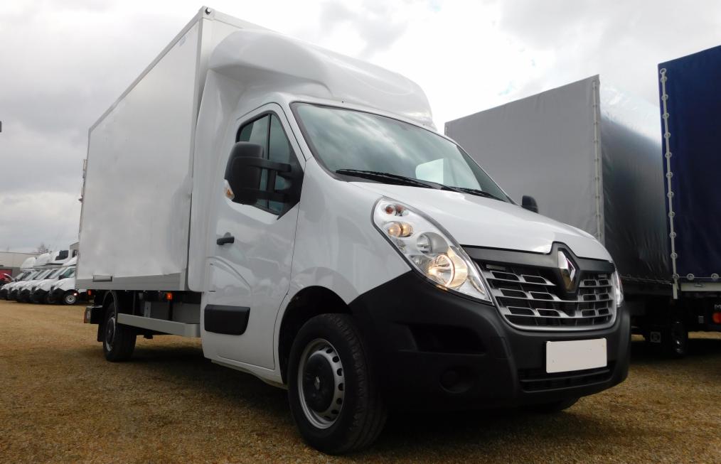 Biały Renault Master z zabudową kontenerową na placu, w tle inne busy i ciężarówki. Widok z przodu, lekko z boku. Światło dzienne, pochmurne niebo.