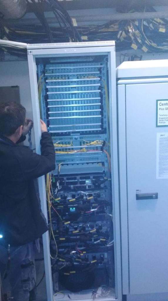 Pracownik w ciemnej bluzie i wzorzystych spodniach manipuluje przy szafie serwerowej z widocznymi kablami i panelami krosowniczymi w pomieszczeniu z instalacjami pod sufitem.