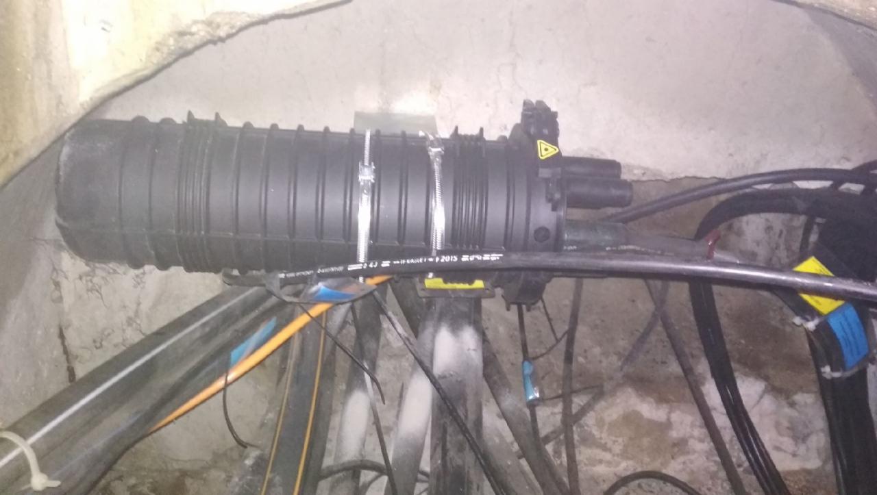 Czarna mufa światłowodowa zamocowana na metalowej konstrukcji wewnątrz betonowego tunelu, widoczne kable i oznaczenia ostrzegawcze.