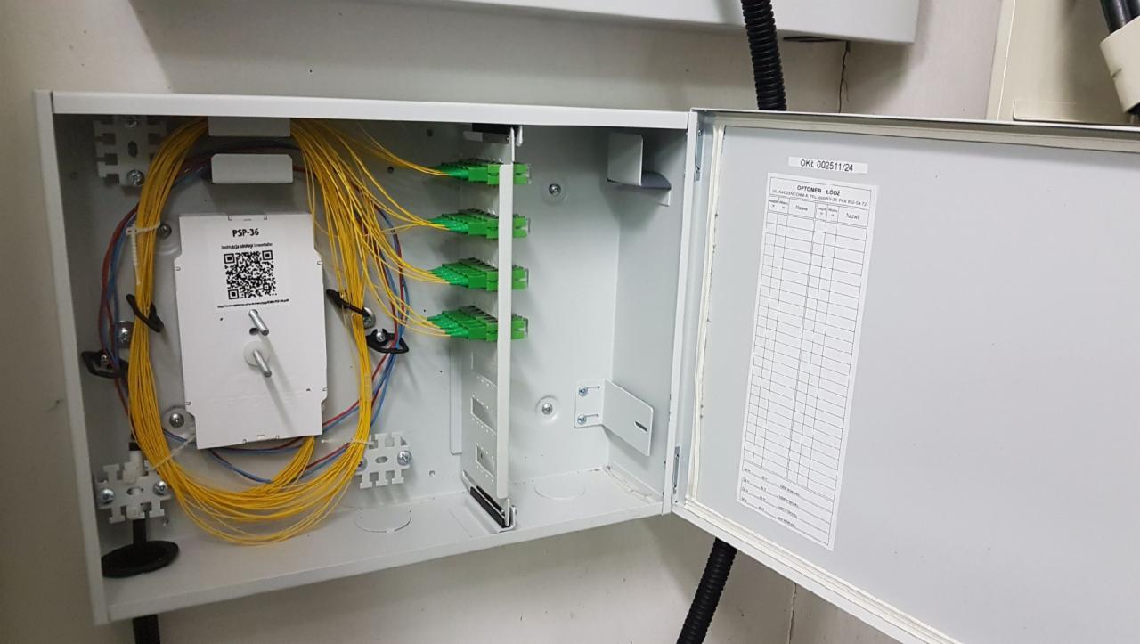 Otwarta skrzynka telekomunikacyjna z żółtymi kablami światłowodowymi, zielonymi złączkami i białą kartką z kodem QR PSP-36