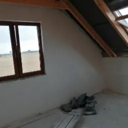 Poddasze po nałożeniu świeżych tynków, widoczne okno dachowe, okno w ścianie szczytowej z widokiem na pole i sterta cegieł na podłodze.