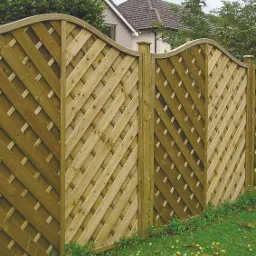 Drewniane, ażurowe ogrodzenie panelowe z łukowatym wykończeniem, widoczne na tle domu w Wołominie, otoczone zielonym trawnikiem z drobnymi żółtymi kwiatami.