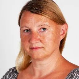 Portret kobiety z blond włosami, w bluzce z geometrycznym wzorem, na jasnym tle.