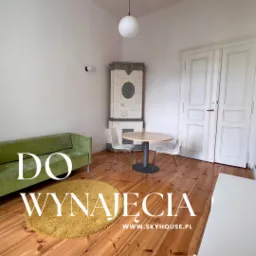 Jasne wnętrze pokoju z zieloną sofą, okrągłym dywanem, piecem kaflowym i okrągłym stołem z krzesłami, drewniana podłoga, informacja o wynajmie na zdjęciu.