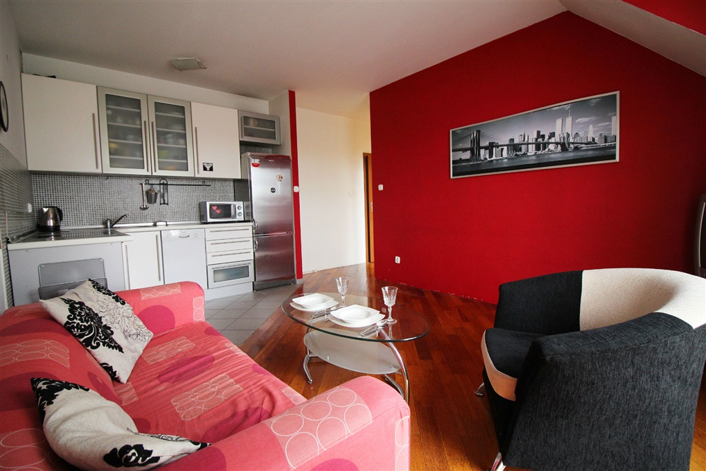 Jasne wnętrze mieszkania z aneksem kuchennym, czerwona ściana z panoramą miasta, różowa sofa z poduszkami i szklany stolik z nakryciem dla dwóch osób.